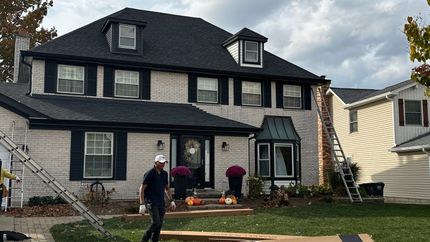 A worker stands in the front yard of a two-story home with a black roof and a ladder propped against the exterior.