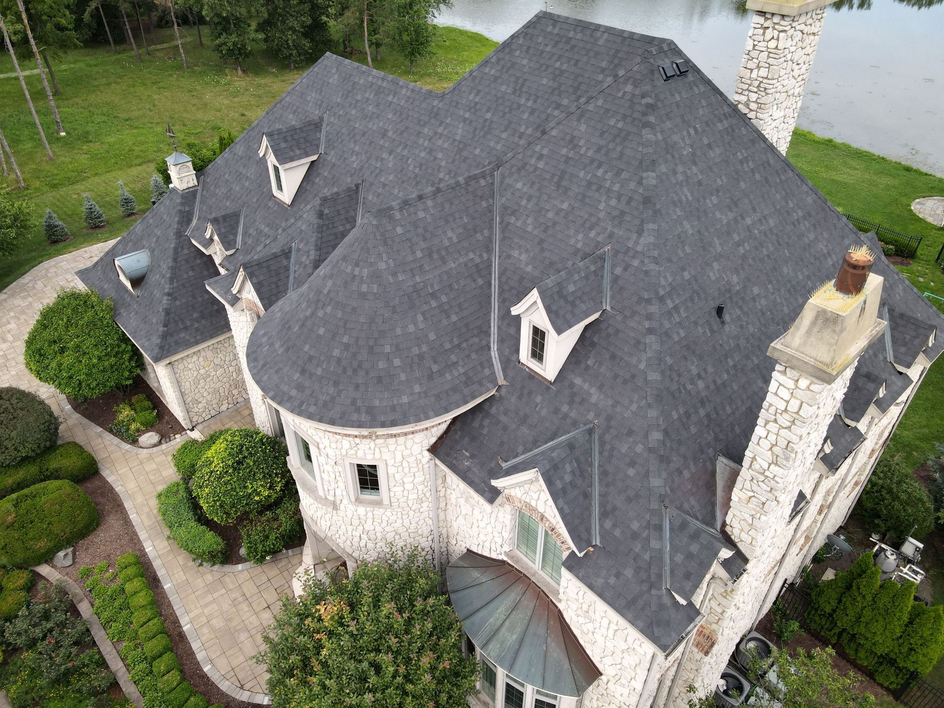 An aerial view of a stone house with a dark shingled roof, a turret, and landscaped grounds next to a body of water.