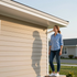 A person in a light blue shirt and jeans stands by the beige siding of a house, looking up at the eaves.