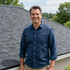 A smiling person in a blue button-down shirt stands in front of a shingled roof with trees in the background.