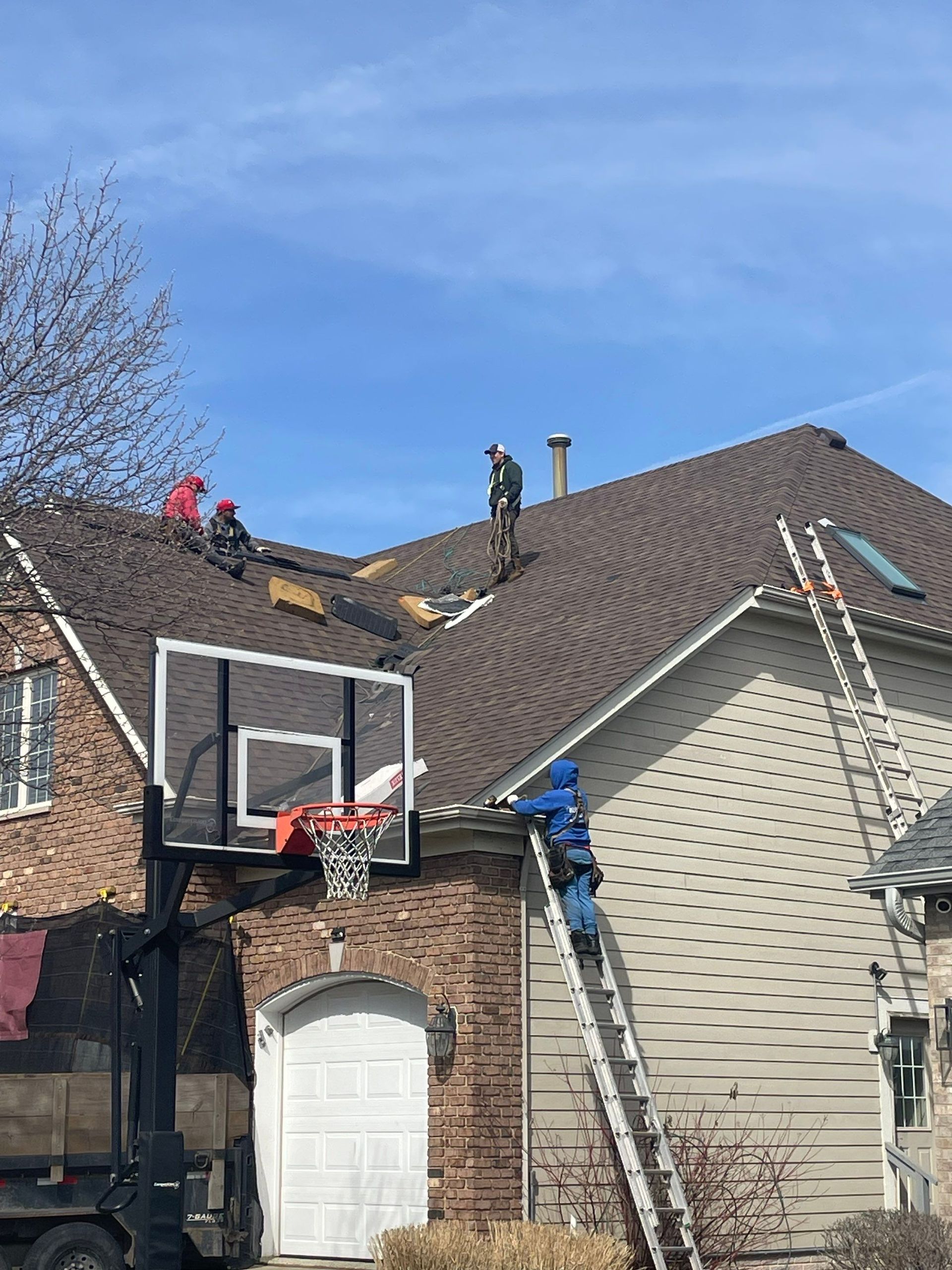 Workers on a residential roof removing shingles, with a ladder leaning against the house and a basketball hoop nearby.