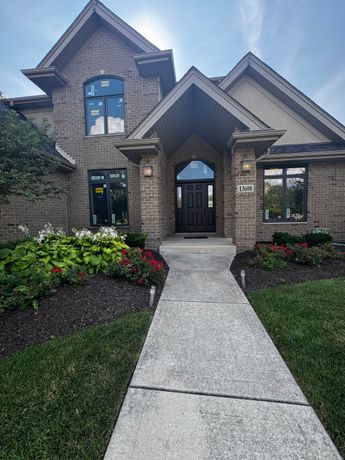 A two-story brick house with a front walkway, dark front door under a peaked portico, and landscaping on a sunny day.