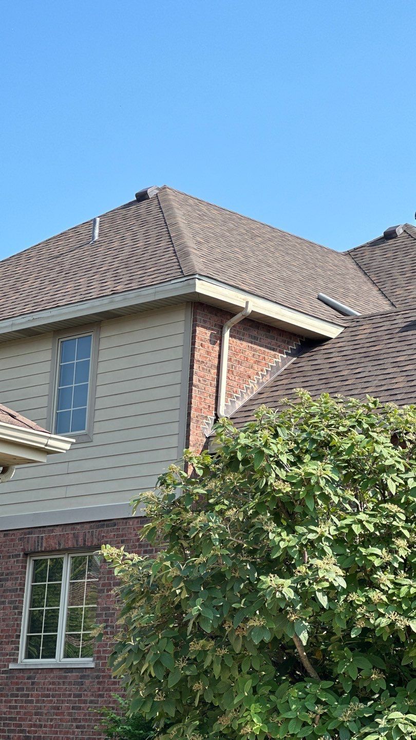 A house exterior featuring light-tan horizontal siding, red brick masonry, gray shingles, and a window partially hidden by.