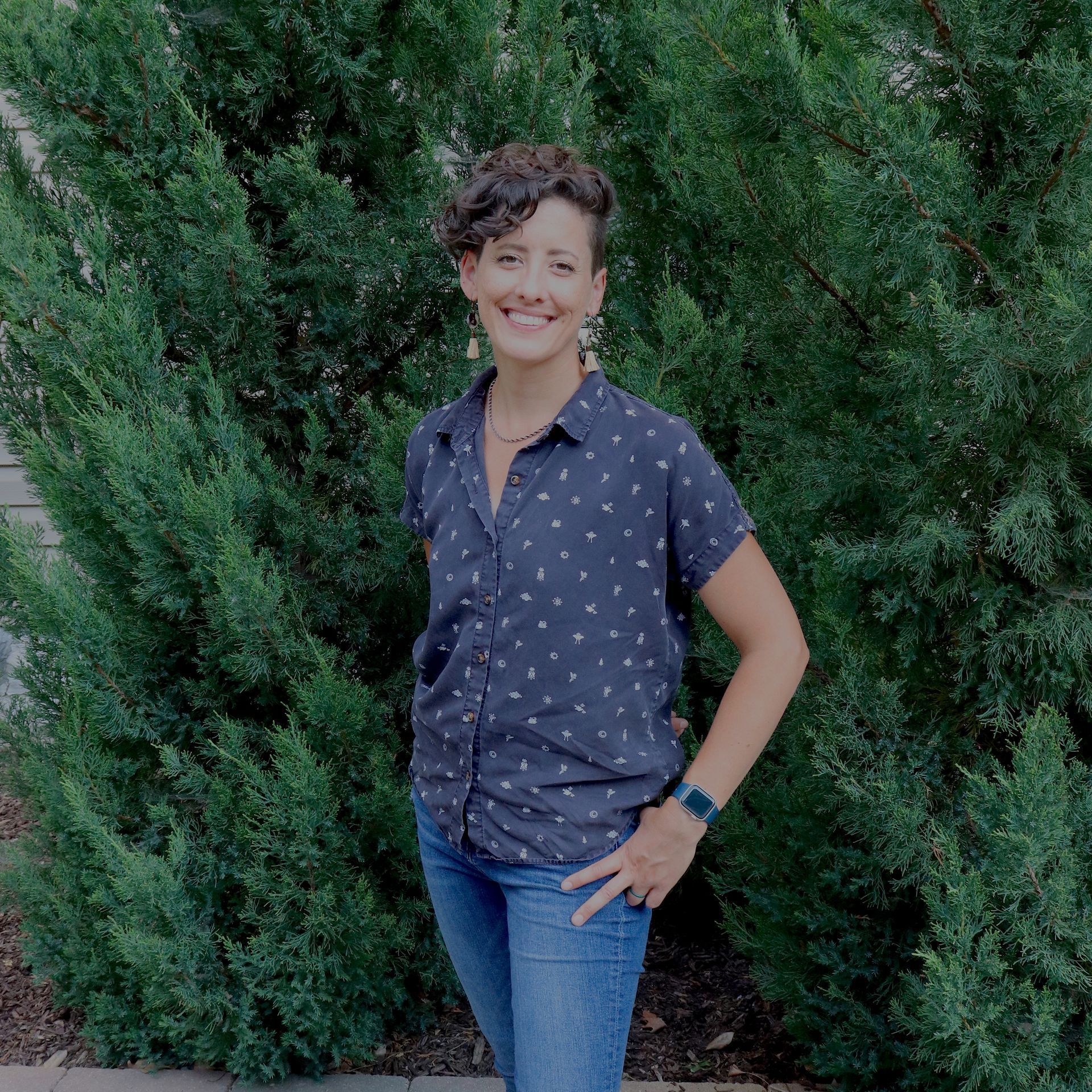 A woman in a blue shirt and jeans is standing in front of a tree.