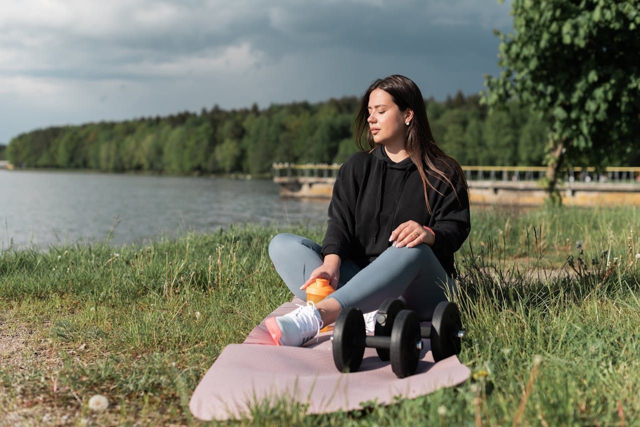 A woman is sitting on a yoga mat next to a lake.