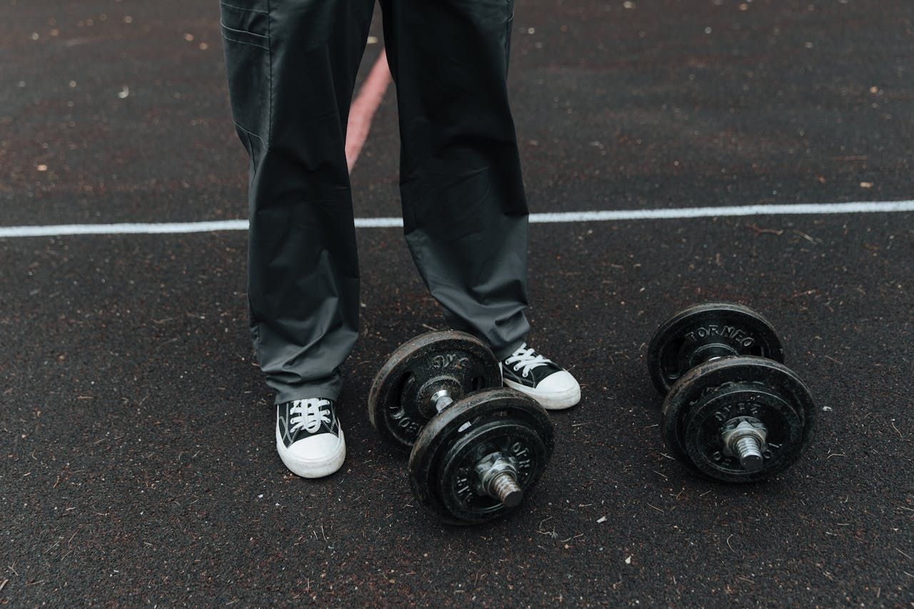 Person standing with black pants and white sneakers near two pairs of dumbbells on a dark surface.