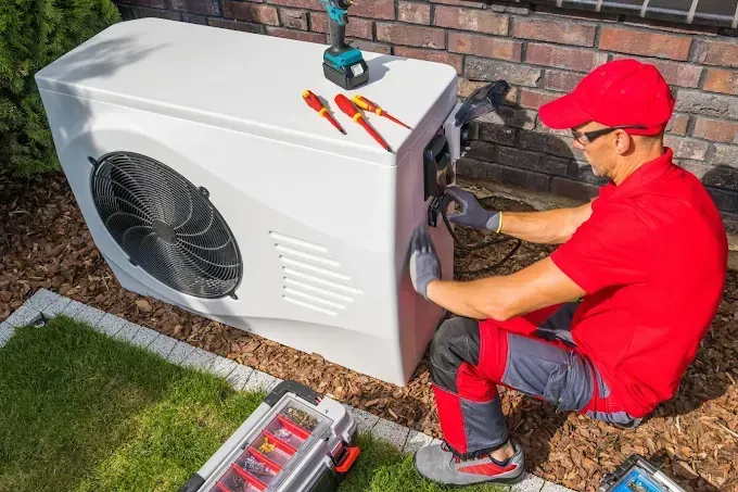 Person in red uniform servicing a white heat pump outdoors with tools.