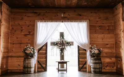 Rustic wooden wedding venue with white drapes, floral arrangements, and a small table at the altar.