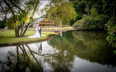 Bride and groom by a pond near a rustic lodge, reflected in calm water and surrounded by trees