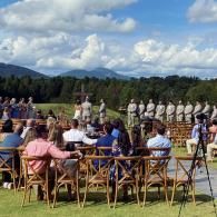Outdoor ceremony with seated guests, uniformed participants, and mountains under a cloudy sky