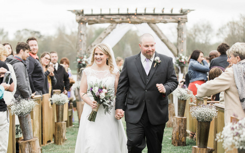 Bride and groom walking down an outdoor aisle under a wooden arch, guests cheering on both sides