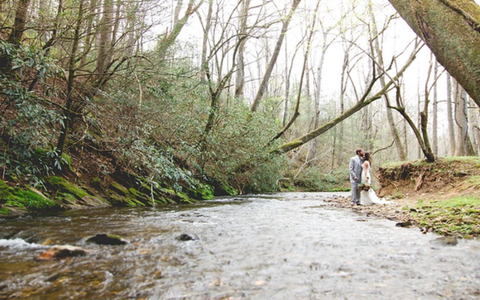 Two people stand by a shallow creek in a wooded forest clearing