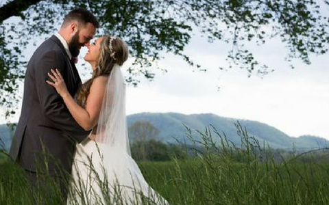 Wedding couple embracing in a grassy field with mountains and trees in the background