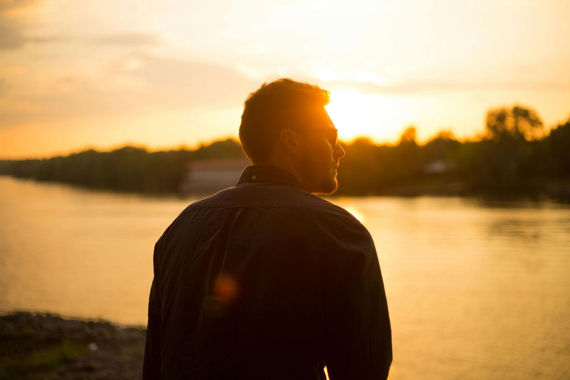 A man is standing on the shore of a lake at sunset.
