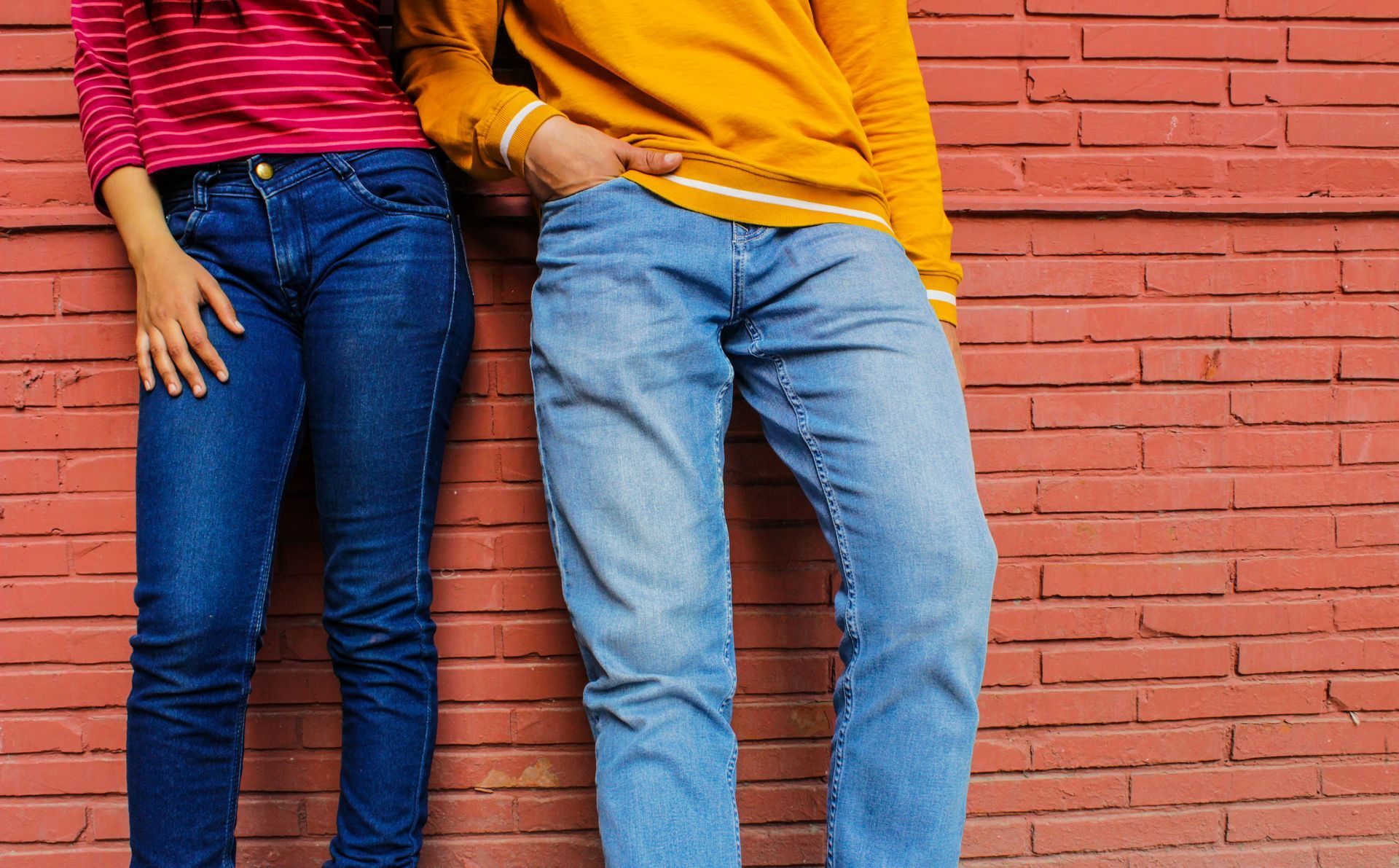 Two people lean against a red brick wall. One wears a pink striped shirt and jeans, the other a yellow sweatshirt and jeans.