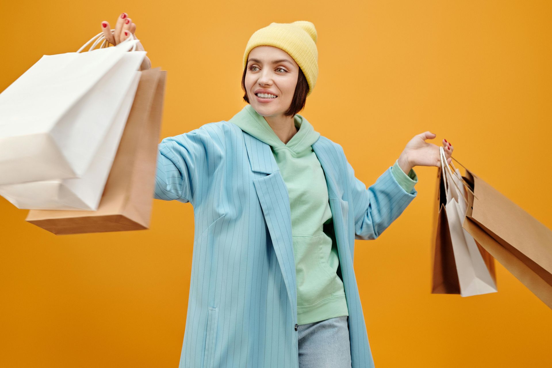 A woman in a blue coat and yellow hat is holding shopping bags.