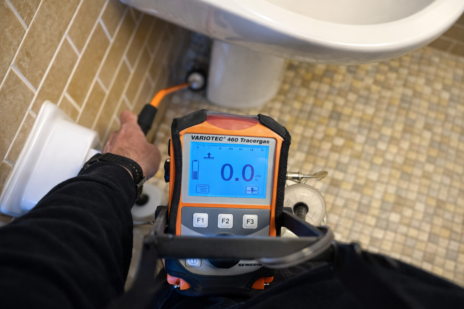 A person holds a hand-held gas detection device near a bathroom sink, with the display reading zero.