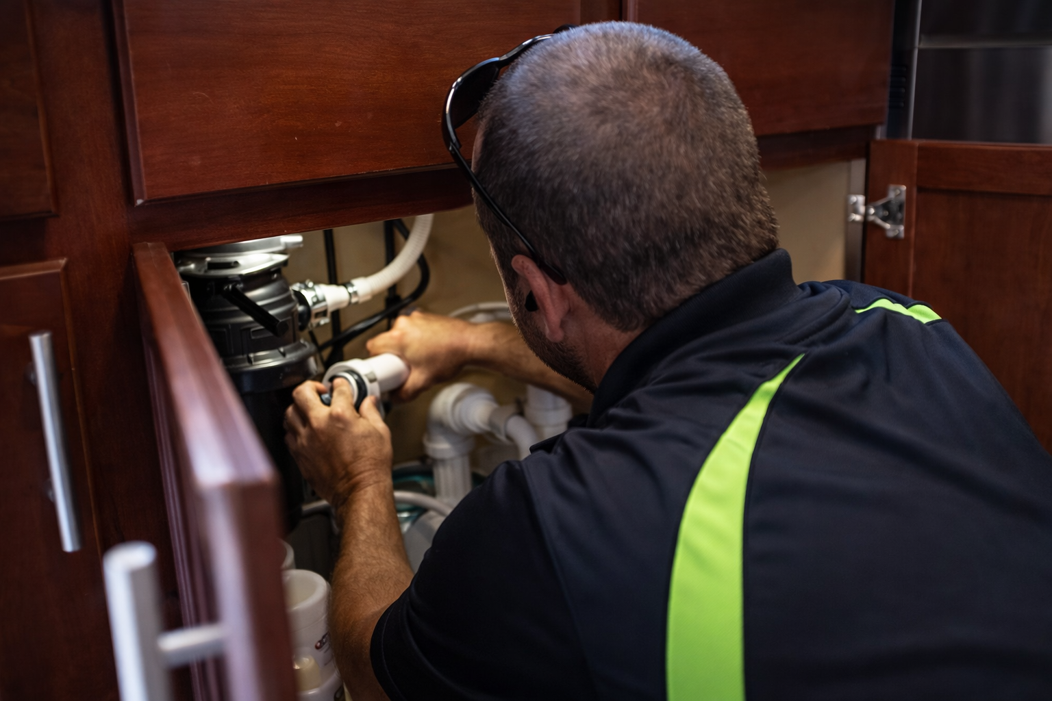 A technician in a black and neon green uniform works on plumbing connections underneath a wooden kitchen cabinet.