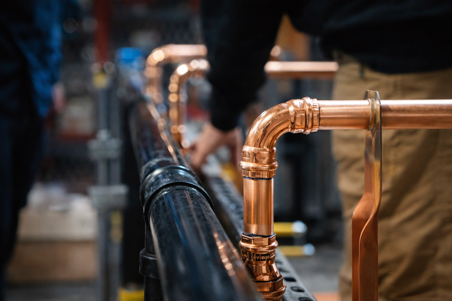Close-up of a worker's hands assembling copper pipes alongside a black metal railing in an industrial setting.