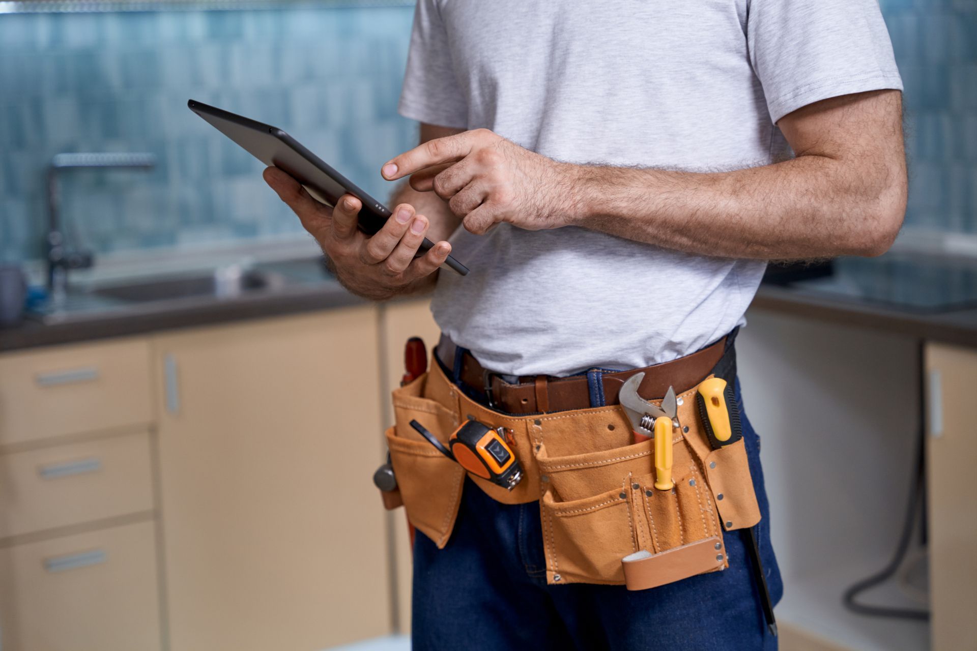 A man wearing a tool belt is using a tablet in a kitchen.