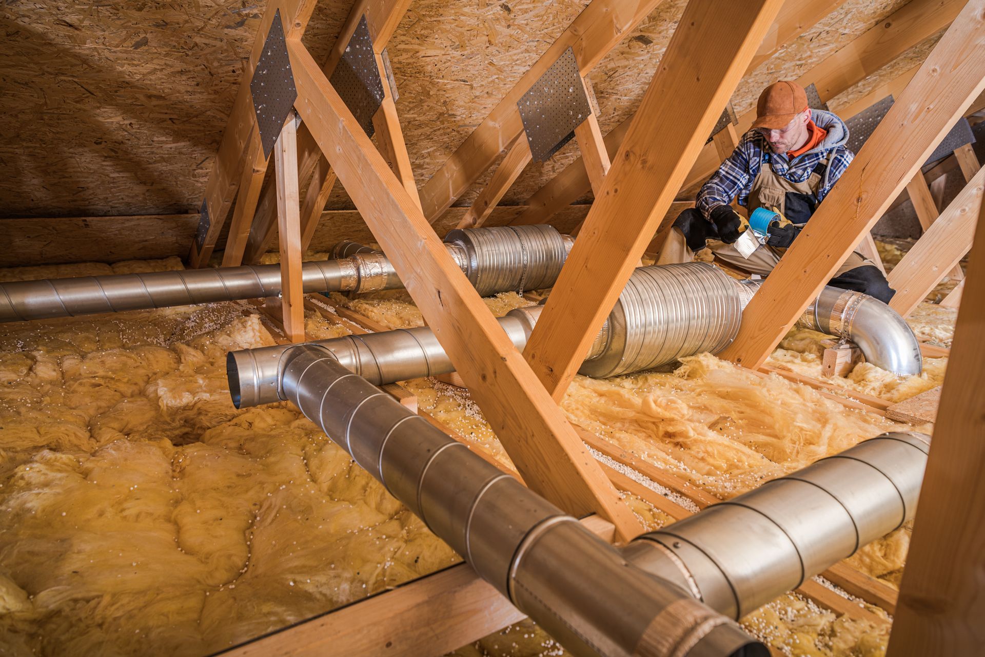 Attic with metal ductwork and wooden beams, with a person sitting among insulation near the top right.