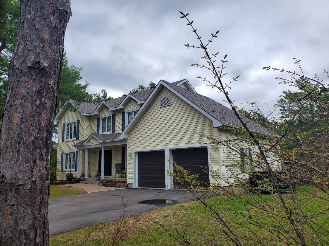 Une grande maison avec deux garages est entourée d'arbres par temps nuageux.