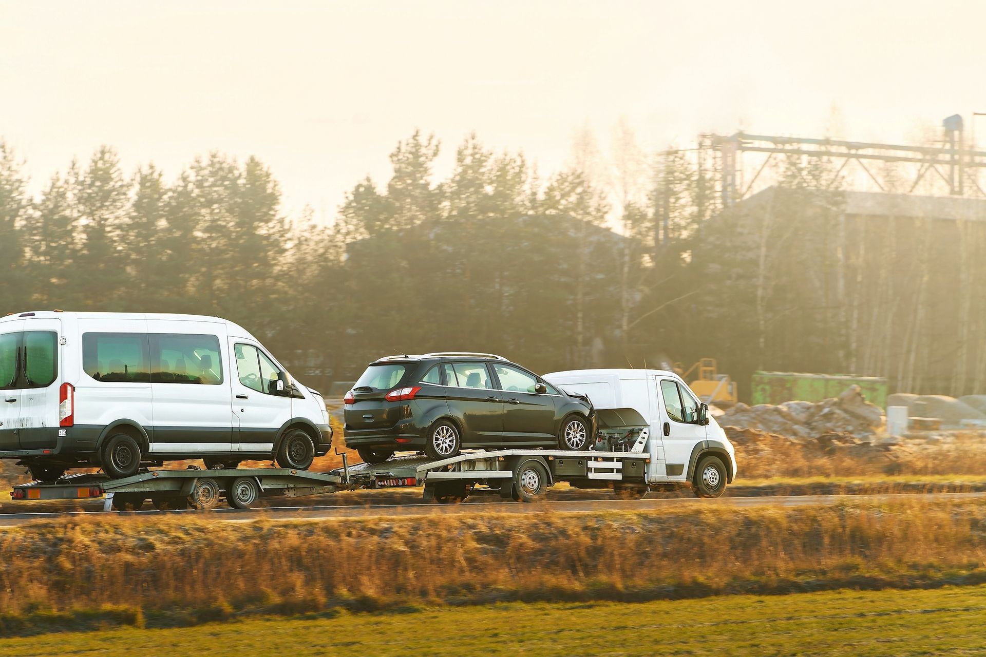 Vehicles towing cars on a rural road. White van and small truck. Brown and green landscape in background.