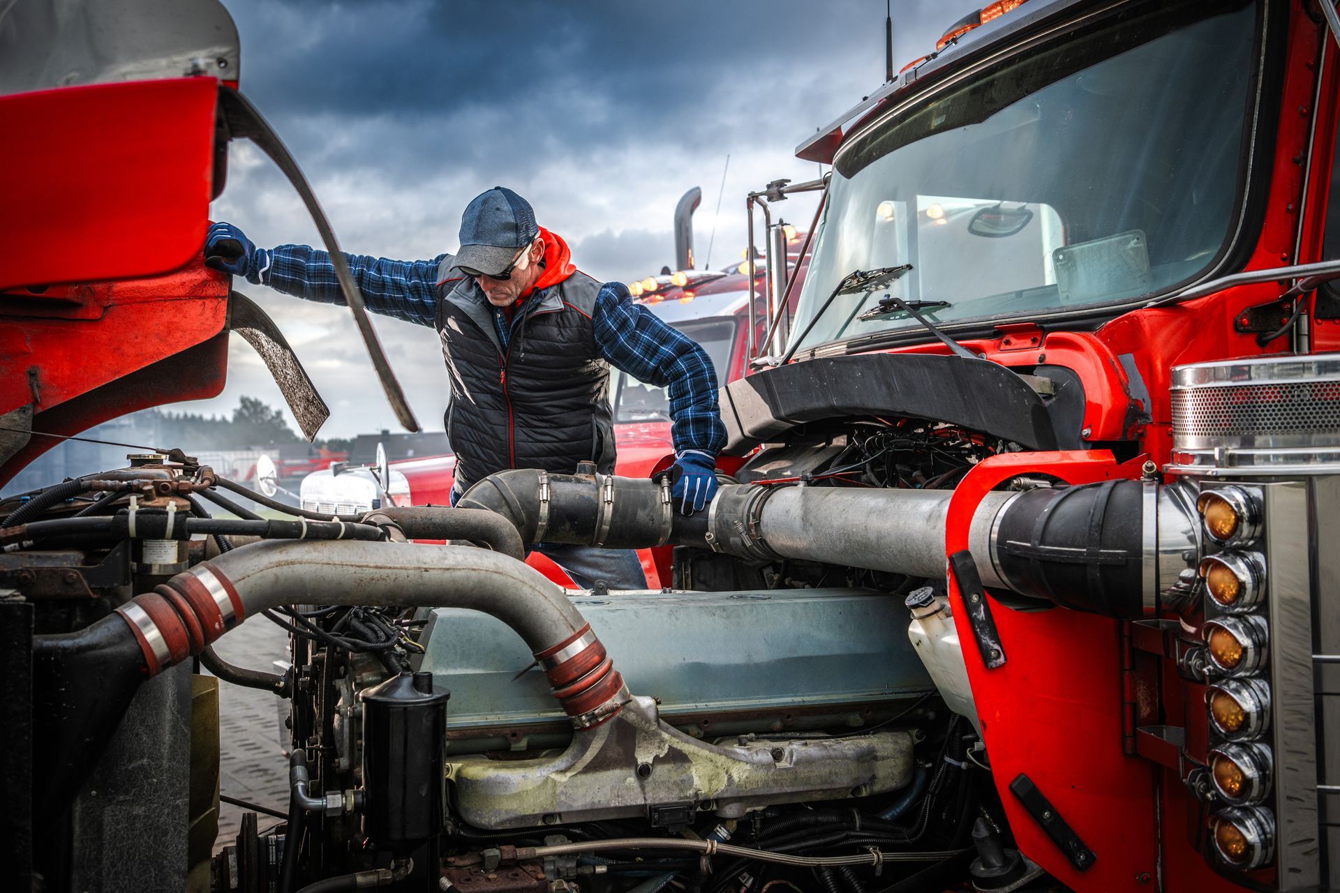 Truck mechanic inspecting the engine of a bright red semi-truck; outdoors, cloudy sky.