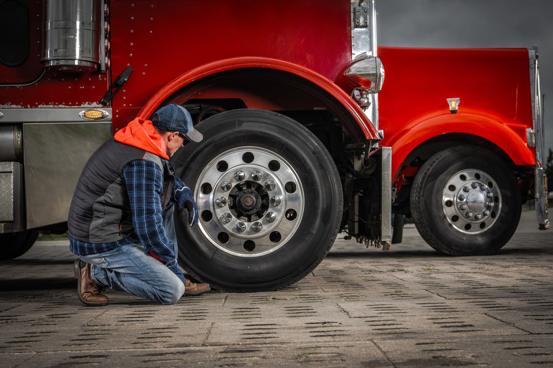 Person kneels to check a semi-truck tire; red truck, brick pavement, overcast sky.