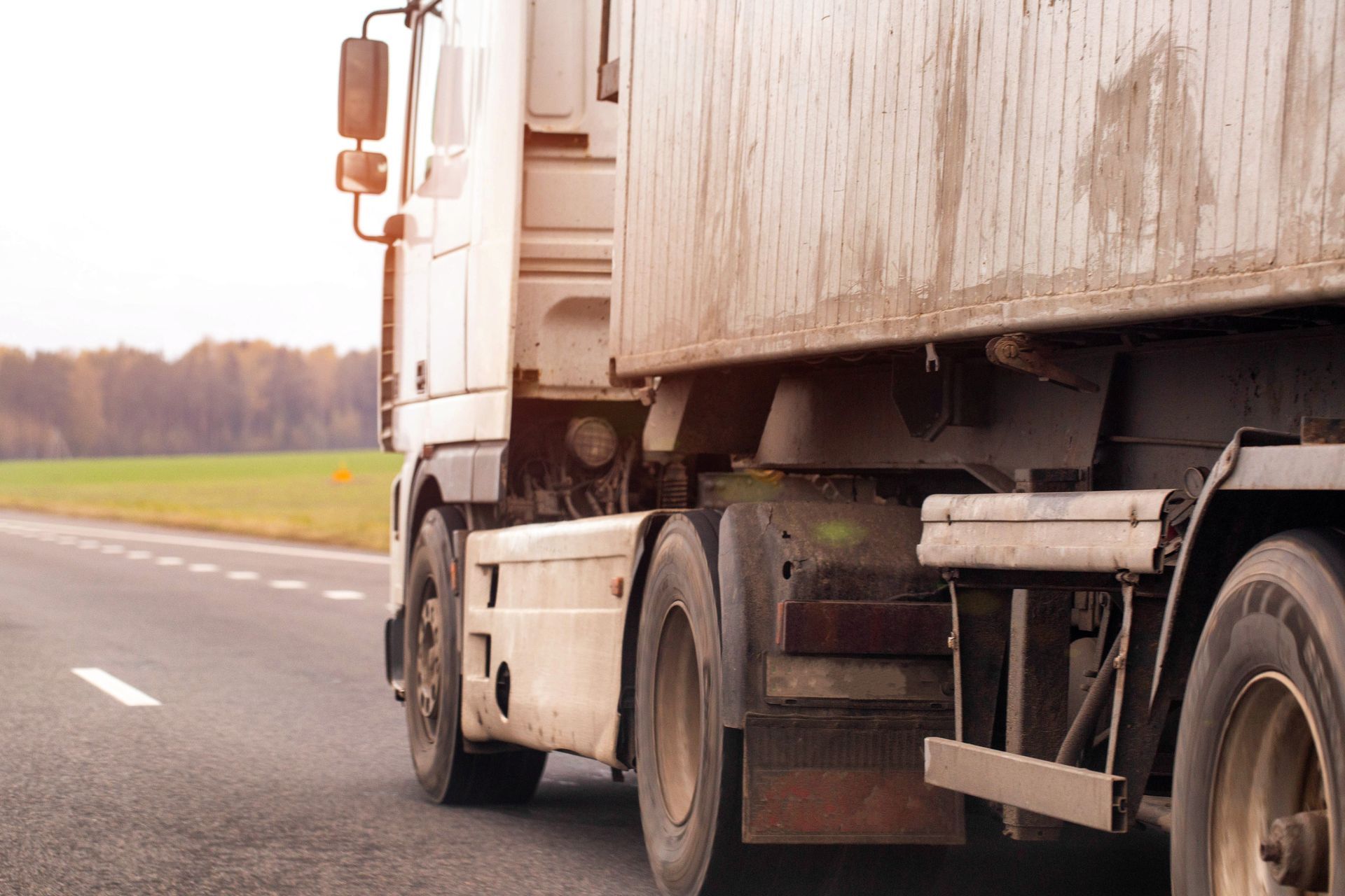 White semi-truck driving on a road with green field and trees in the background.