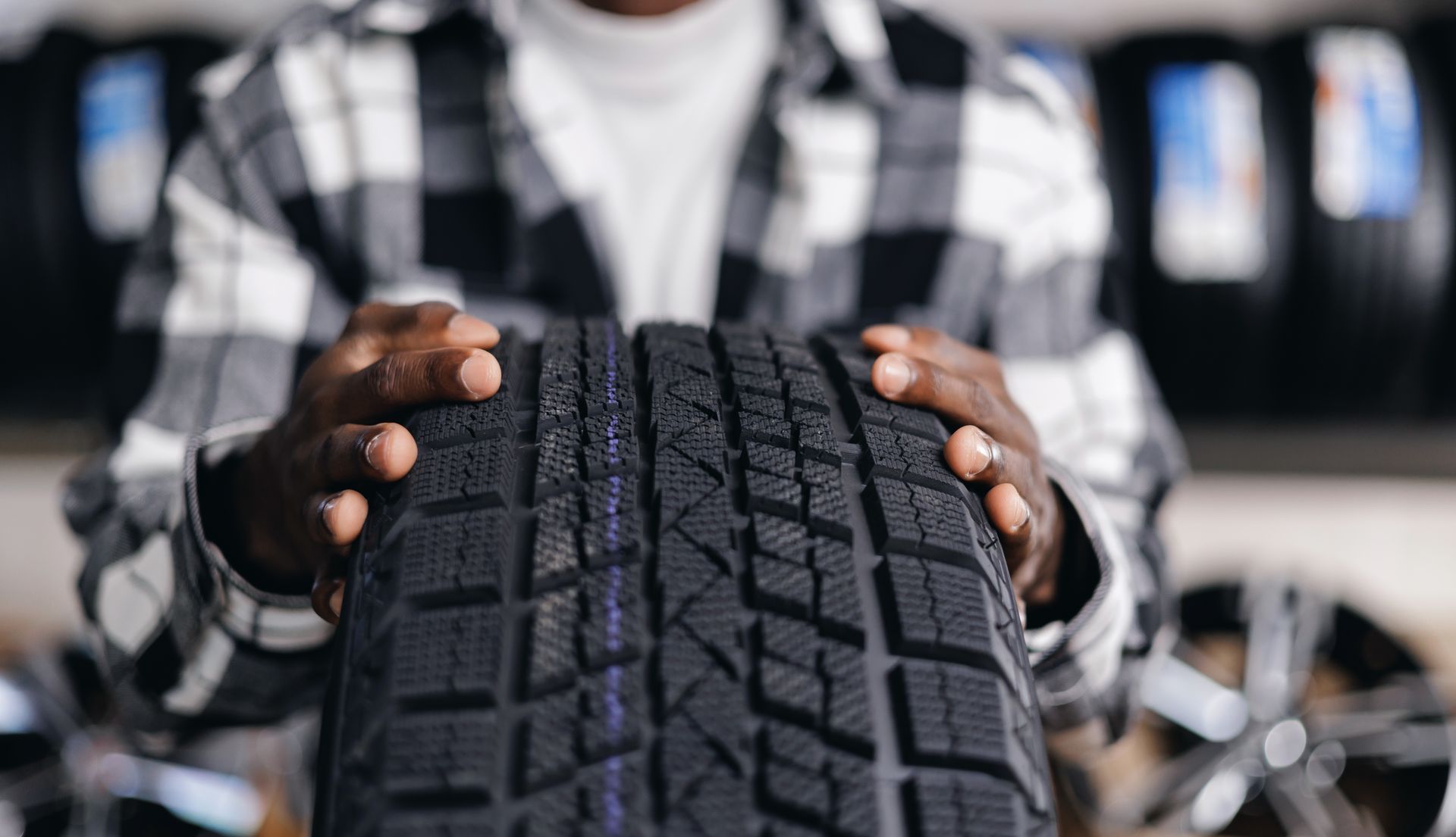 Person holding a tire, showcasing its tread. Tire shop setting, with other tires visible in the background.