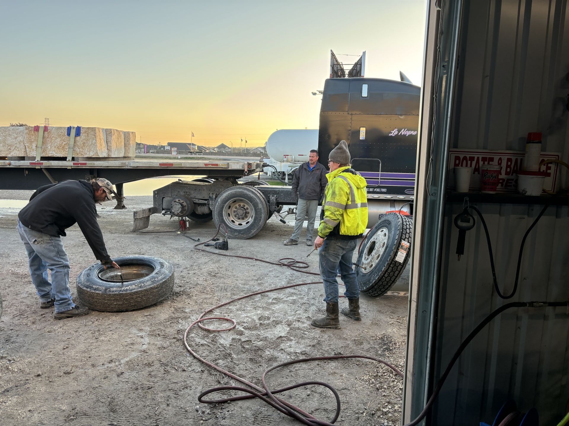 Three people working on a truck tire in a repair bay. One person is handling the tire while others observe.
