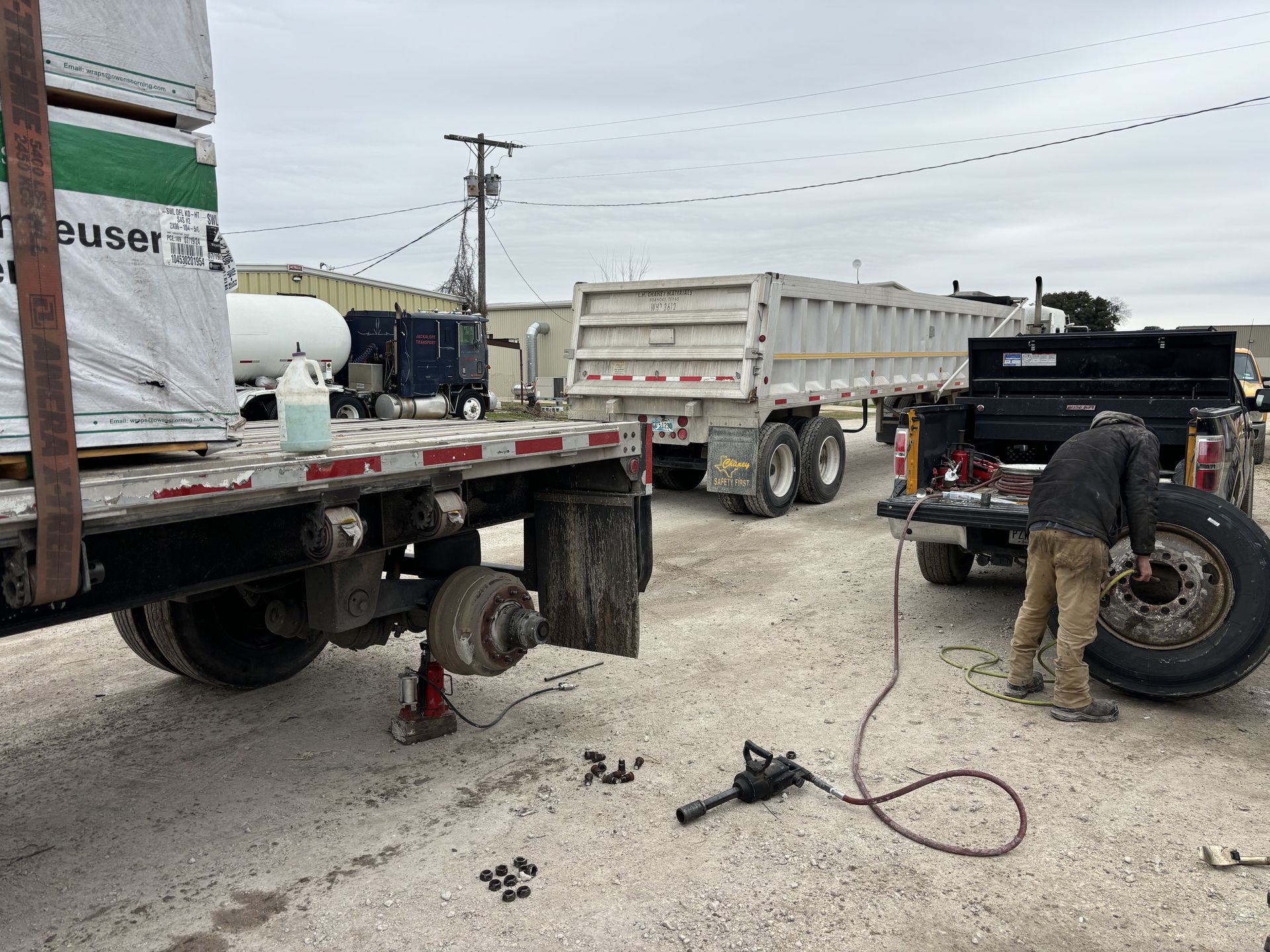A person changes a tire on a flatbed trailer, another trailer and truck are nearby, on a cloudy day.