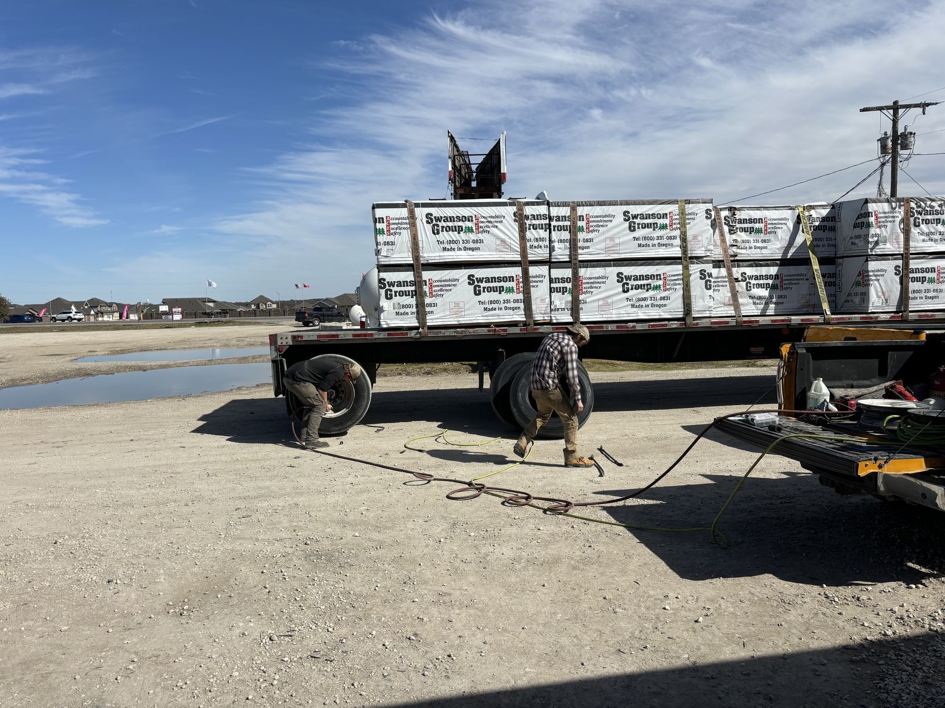 Flatbed trailer loaded with lumber being secured by workers in a gravel lot on a sunny day.