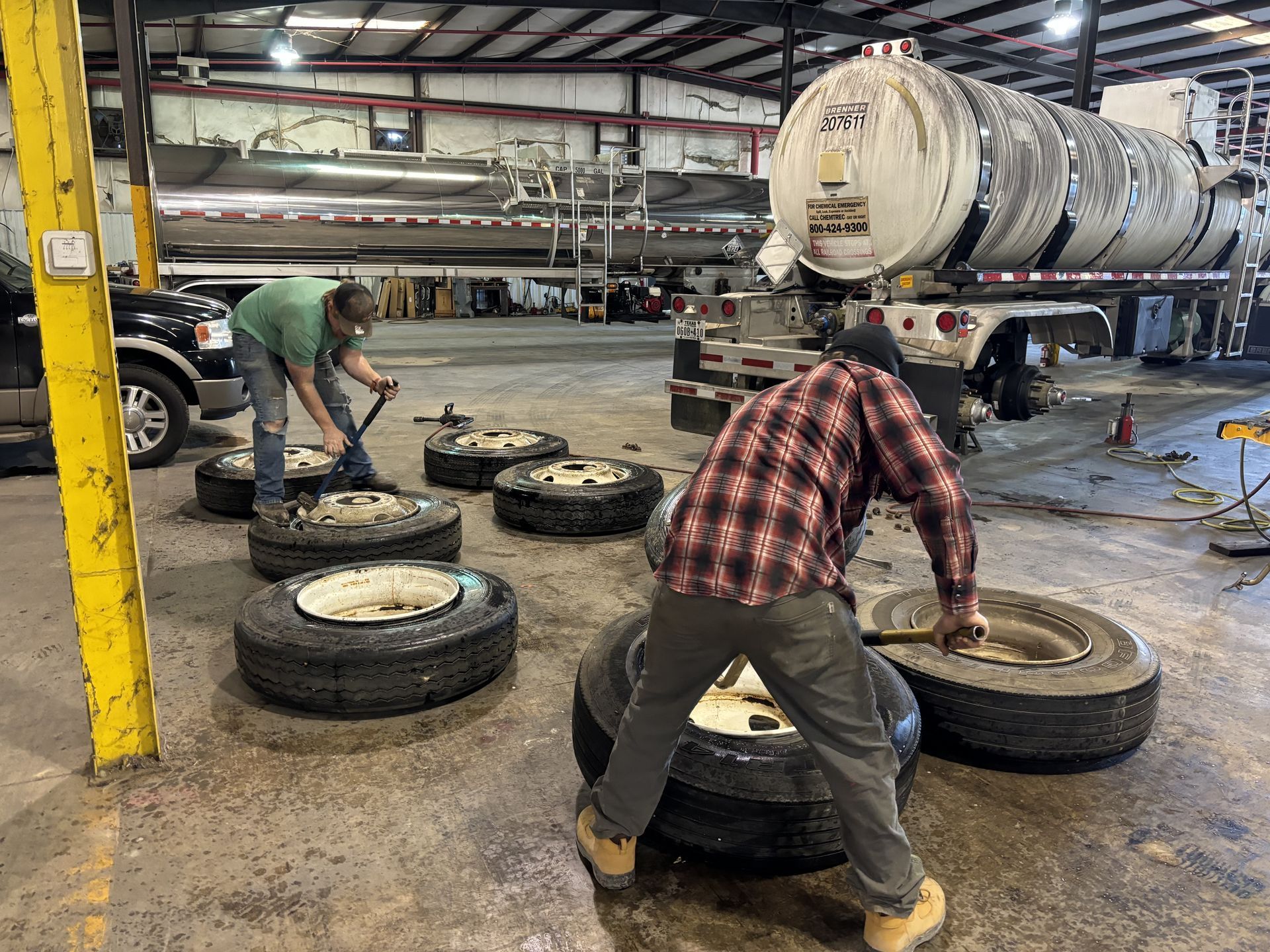 Two workers changing truck tires in a shop with a tanker truck in the background.