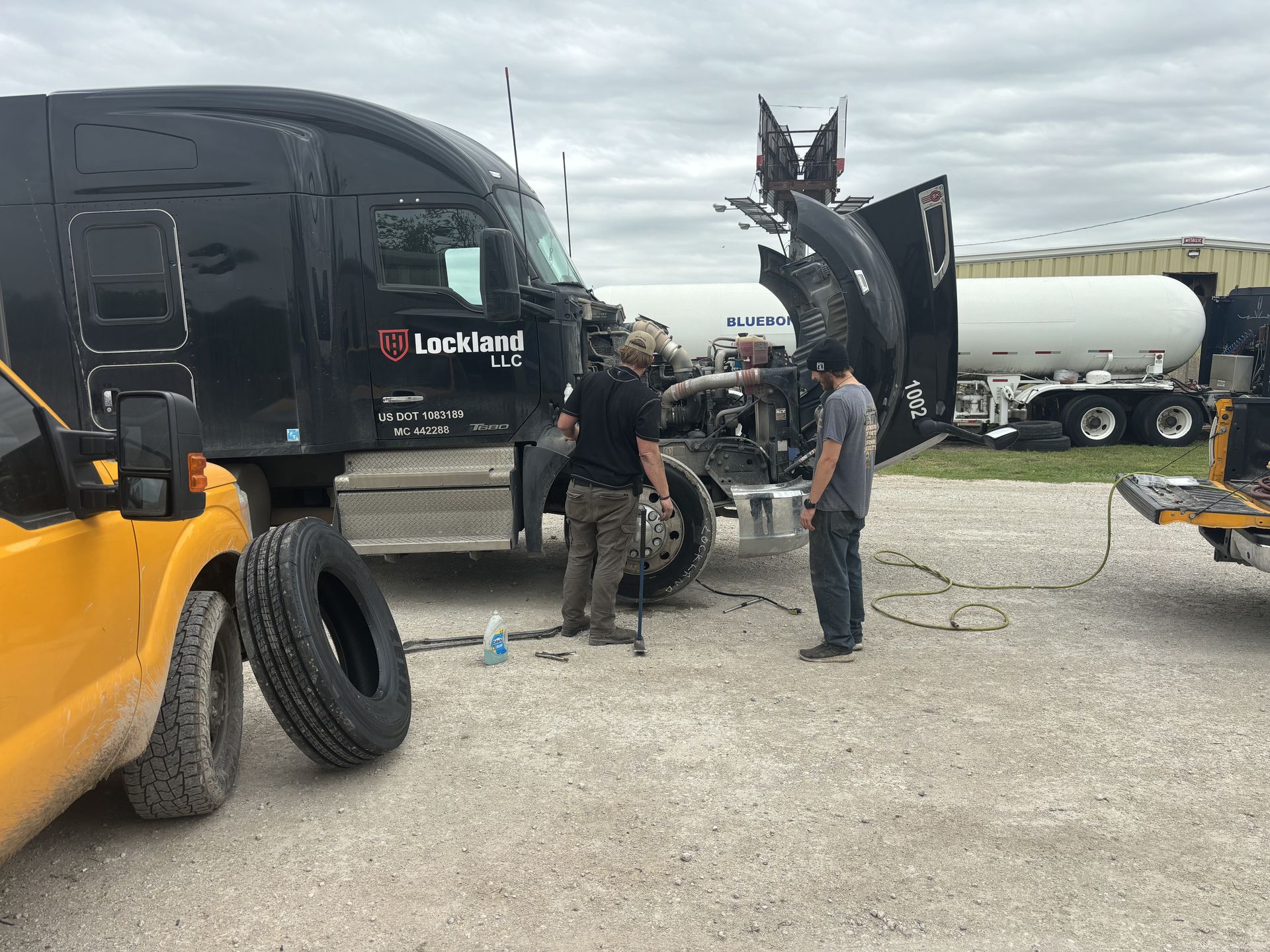 Two people working on a black semi-truck with open hood. Yellow vehicle, spare tire, and propane tank visible in a gravel lot.