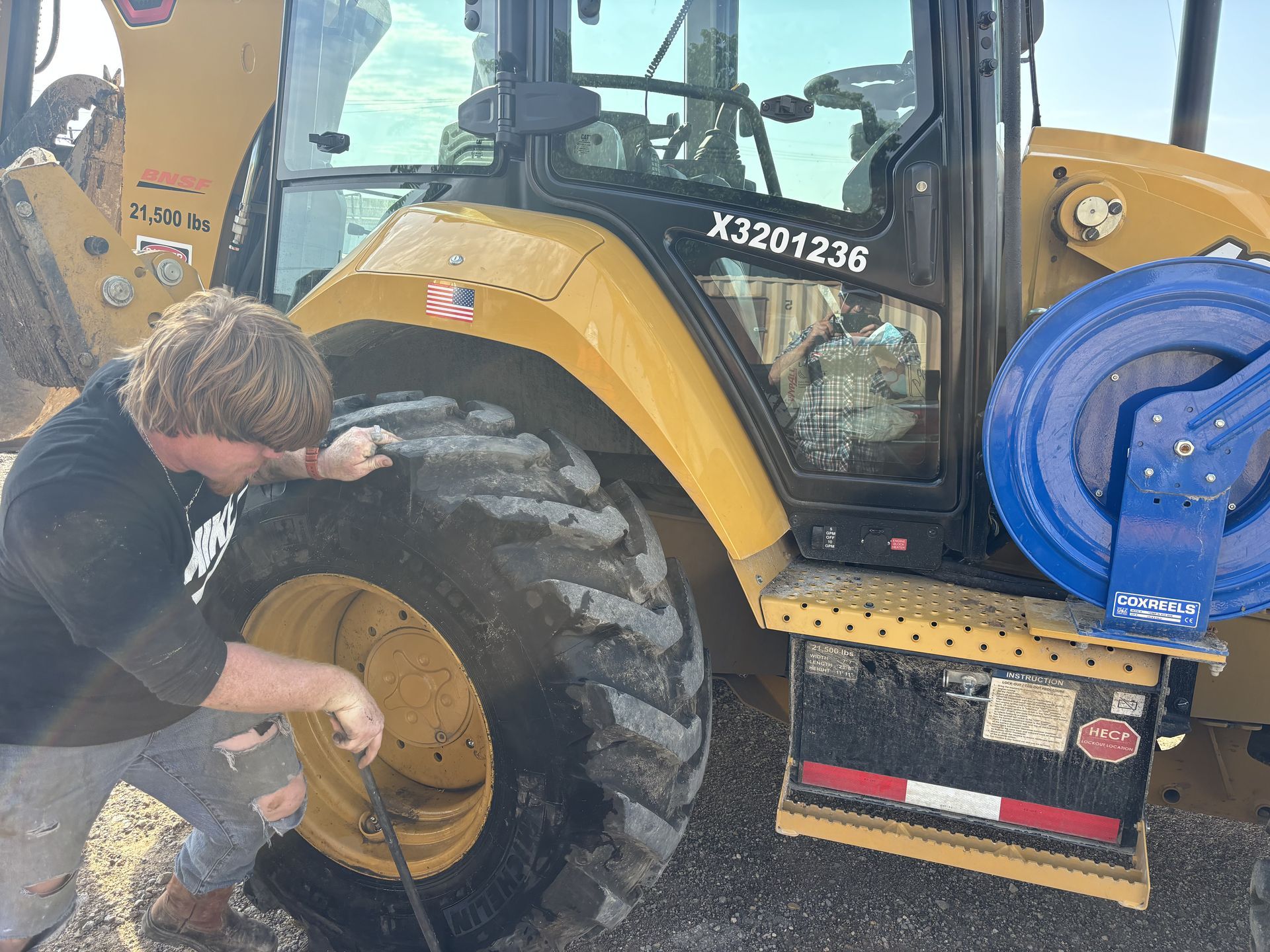 Man checks the tire of a yellow Caterpillar backhoe. The backhoe has a blue attached element and the man is wearing a black t-shirt.