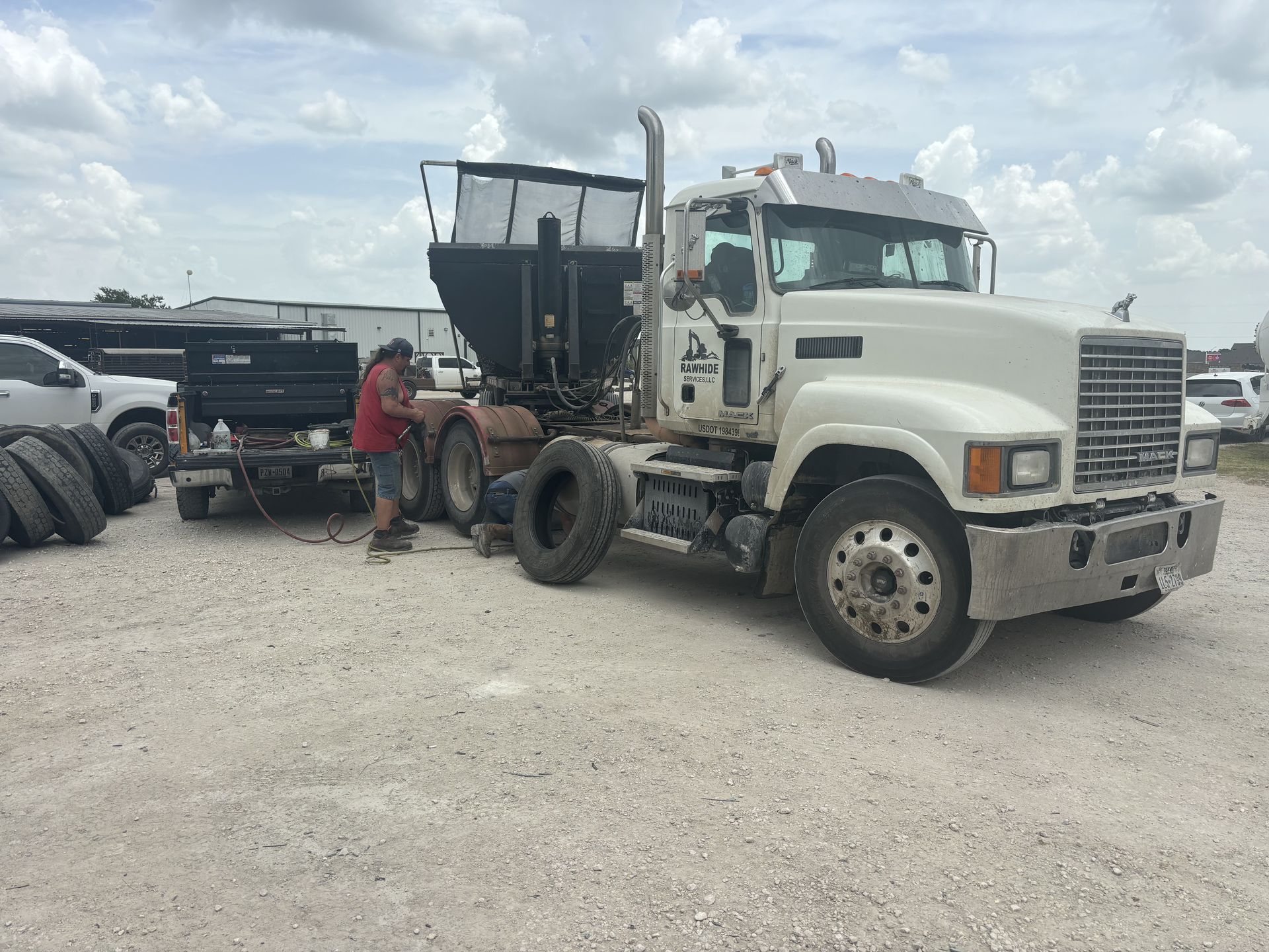 White Mack semi-truck with a black trailer; a person works on the truck in a gravel lot on a cloudy day.