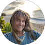 Smiling woman in a blue shirt and necklace standing outdoors by a coastal overlook.