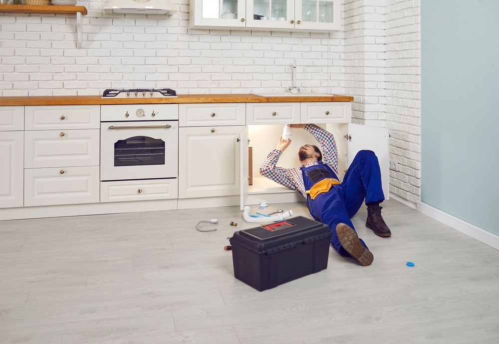 A plumber in blue work coveralls works under a kitchen sink, with a toolbox placed on the floor nearby.