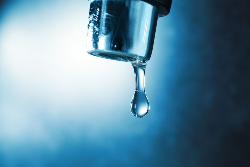A close-up of a metal faucet tip with a single, clear water droplet suspended, set against a dark blue background.