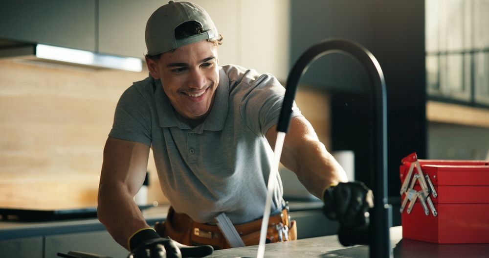 A smiling plumber in a grey polo and baseball cap repairs a modern kitchen faucet, with a red toolbox on the counter.