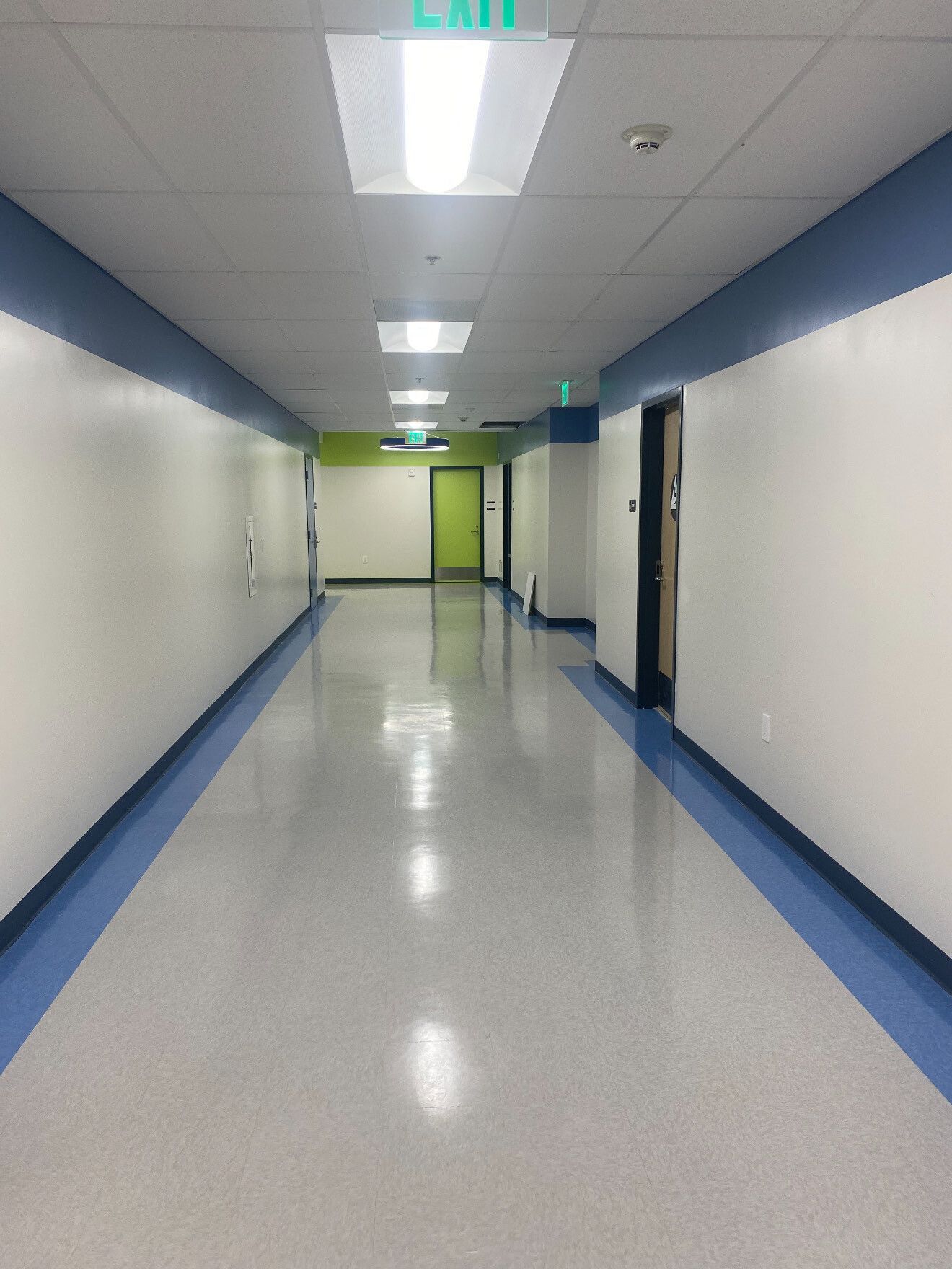 Long hallway with white walls, blue trim, and shiny floors. Green and white exit signs are visible.