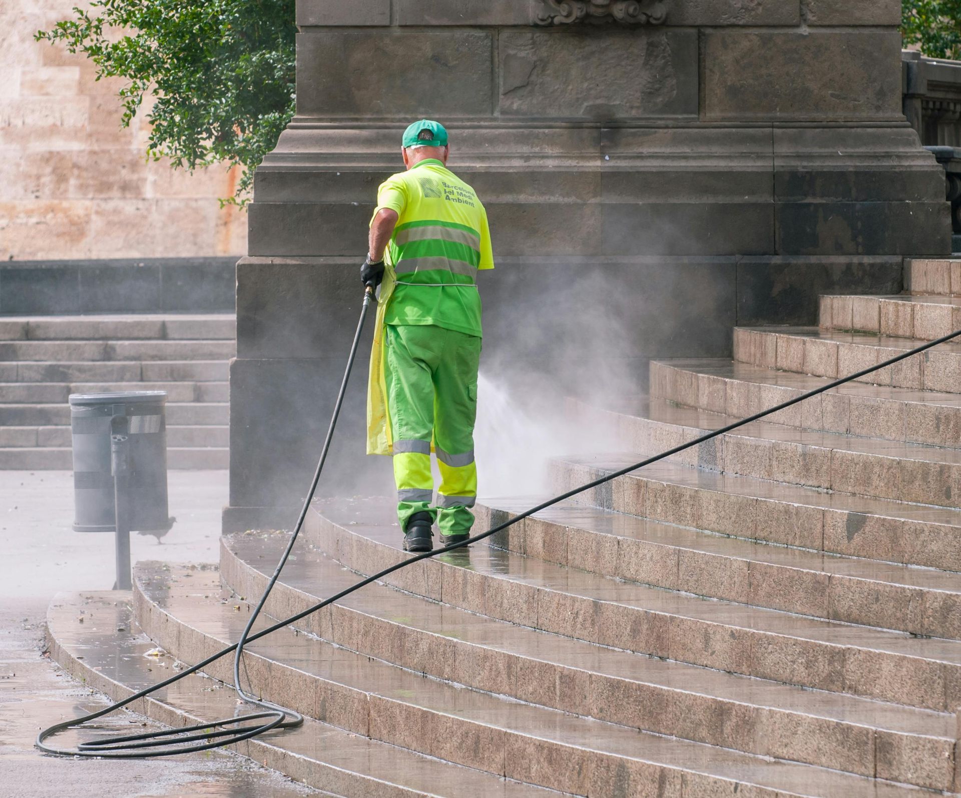 A worker in neon green uniform pressure washes stone steps, generating a mist of water.