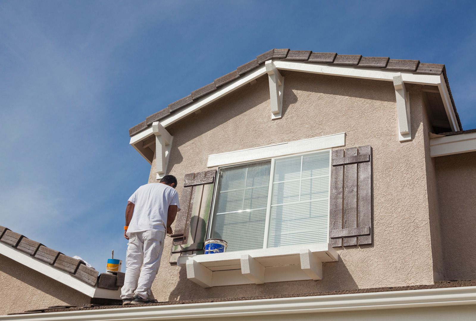 Person painting the exterior of a two-story house with shutters. The sky is blue.