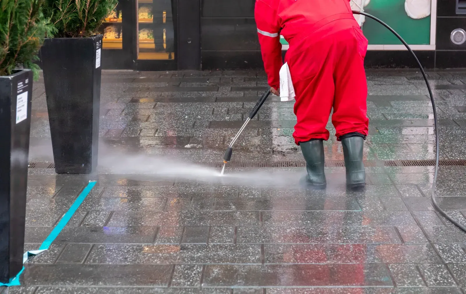 Person in red coveralls power-washing a wet brick sidewalk.