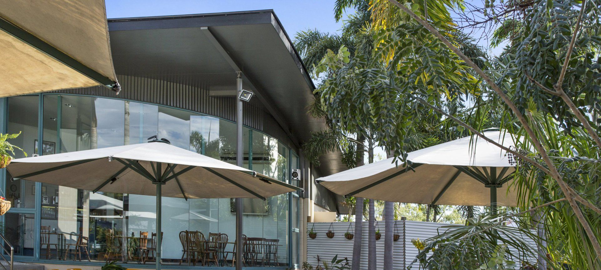 Outdoor Patio With Umbrellas Near A Building With Glass Walls, Surrounded By Trees — Capricorn Motel & Conference Centre In Parkhurst, QLD