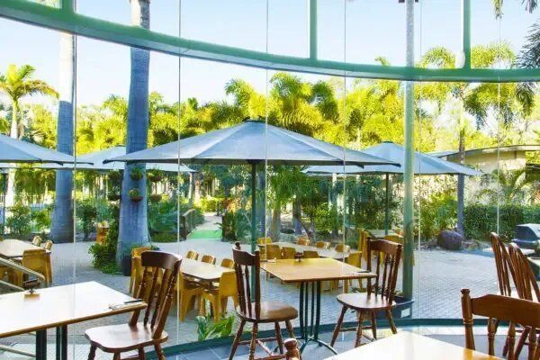 Restaurant Interior With Tables, Chairs, And Glass Walls Overlooking A Green Outdoor Area With Trees — Capricorn Motel & Conference Centre In Parkhurst, QLD