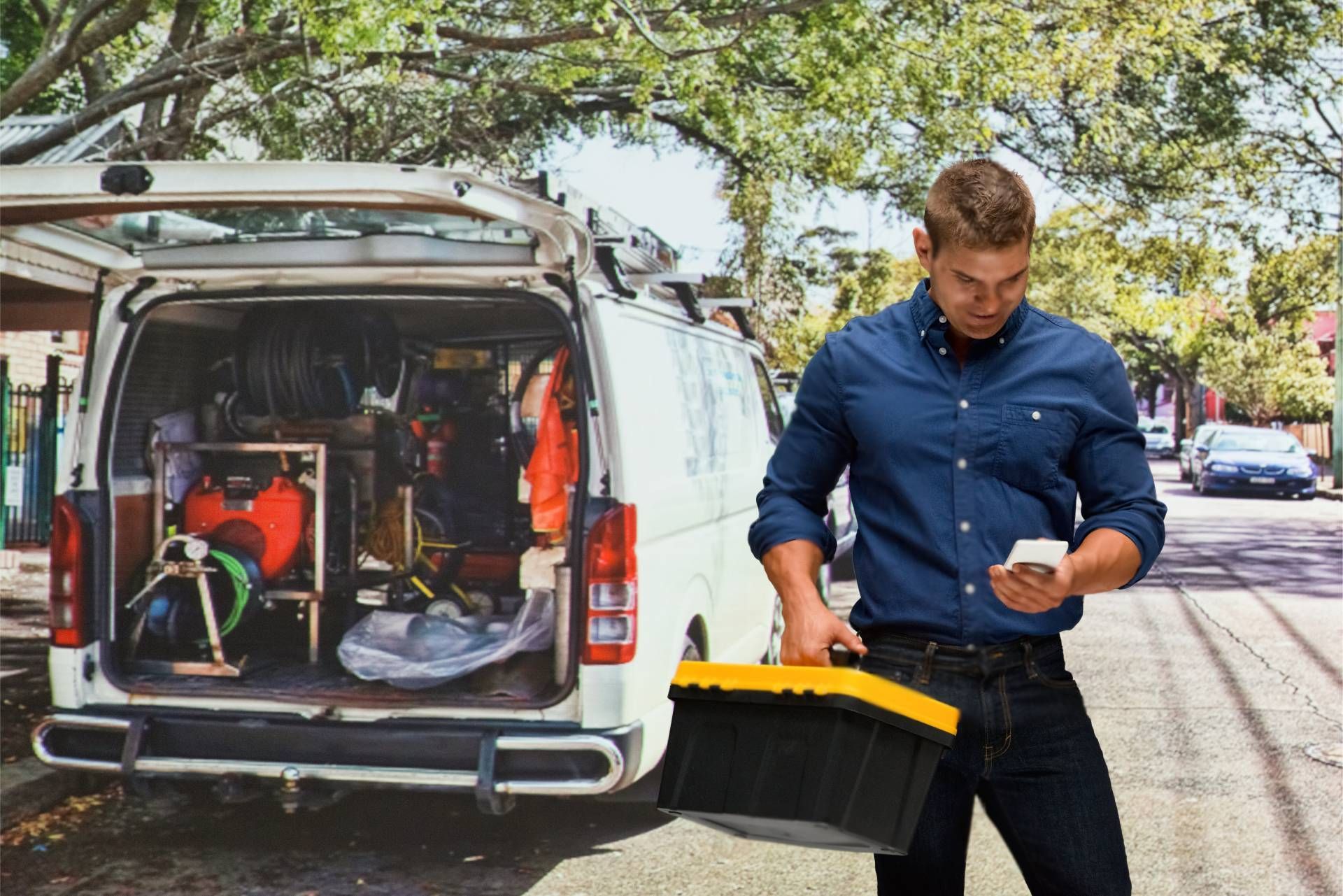 A man is standing in front of a van holding a toolbox and looking at his phone.
