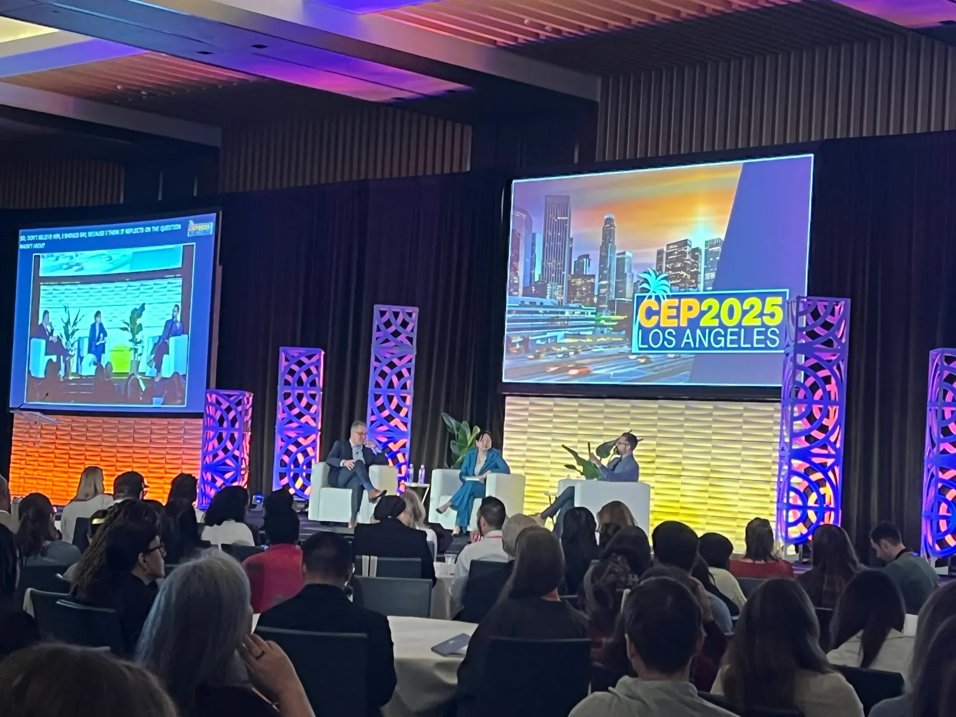 Conference in Los Angeles with speakers on stage, large screens, and an audience in a large ballroom.
